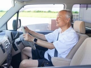 An older man is driving a car with his hands on the wheel. He is smiling and he is enjoying the ride