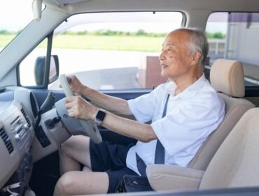 An older man is driving a car with his hands on the wheel. He is smiling and he is enjoying the ride