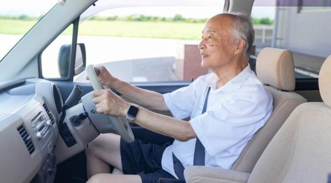 An older man is driving a car with his hands on the wheel. He is smiling and he is enjoying the ride
